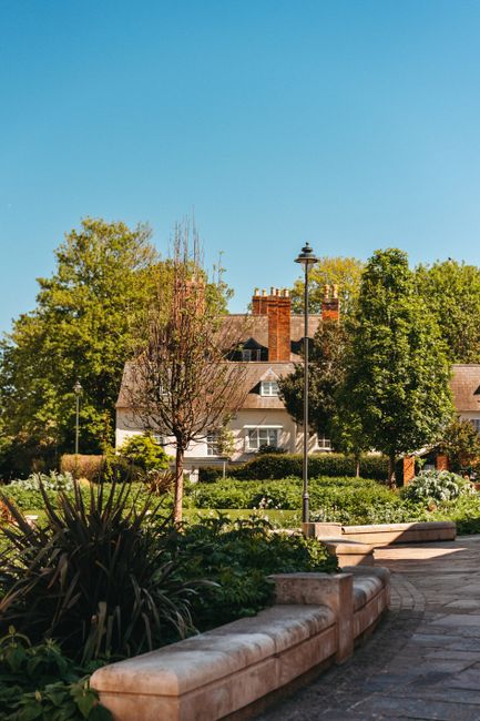 This image depicts a peaceful outdoor scene near Eastcote House Gardens, with a clear blue sky overhead. In the foreground, there is a stone bench with a rounded edge, situated among lush green plants