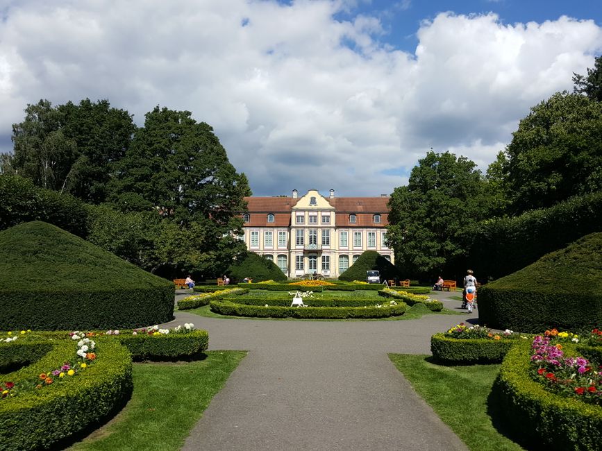 A grand Victorian-style mansion with large windows and a red tiled roof is set behind neatly trimmed topiary and flower beds in a formal garden. The garden features symmetrical, rounded green hedges a